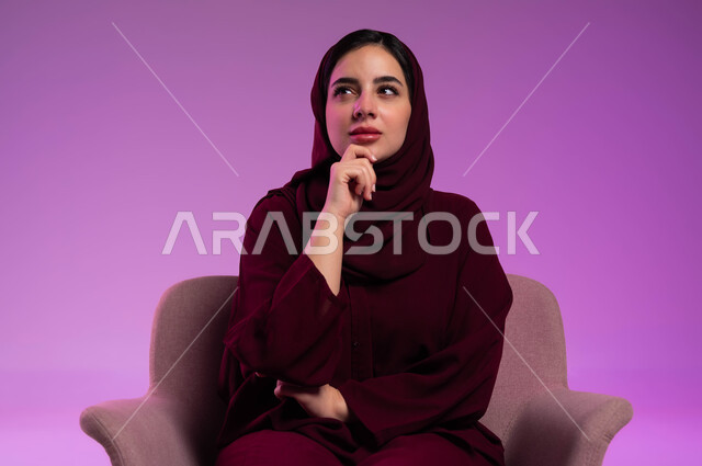 التفكير لأيجاد حل مشكلة ما، بورتريه لامرأة عربيه خليجيه سعوديه محجبه ترتدي العباءة الملونة تجلس على كرسي مريح وتنظر الى الأعلى بإيماءات التمعن والتركيز، شرود الذهن والتأمل والتخطيط للمستقبل، مواجهة المشكلات والتحديات في الحياة العملية، خلفية ملونة