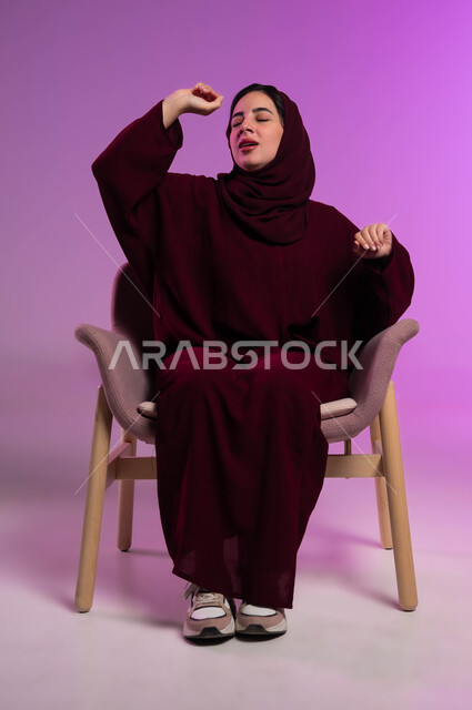 رفع اليدين للأعلى بإيماءات التثاؤب، بورتريه لامرأة عربيه خليجيه سعوديه محجبه ترتدي العباءة الملونة تجلس على كرسي مريح  بتعابير الخمول و الشعور بالنعاس والحاجه الى الراحة و النوم، خلفية ملونة .