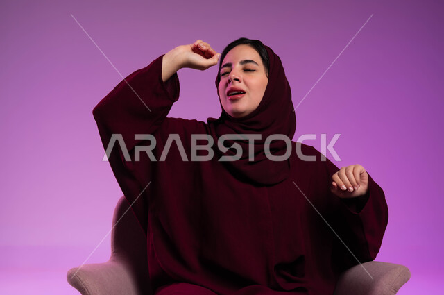 تعابير الخمول و الشعور بالنعاس والحاجه الى الراحة و النوم، بورتريه مقرب لامرأة عربيه خليجيه سعوديه محجبه ترتدي العباءة الملونة تجلس على كرسي مريح ترفع يديها للأعلى بإيماءات التثاؤب، خلفية ملونة .
