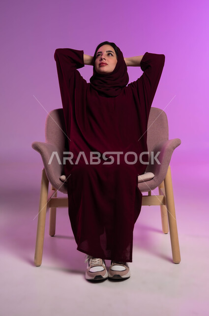 النظر للأعلى إلى شيء ما، شرود الذهن و التمعن ،الشعور بالتعب و الخمول والحاجة إلى النوم, بورتريه مقرب لامرأة عربيه خليجيه سعوديه محجبه ترتدي العباءة الملونة تجلس على كرسي مريح تضع يديها وراء رأسها بتعابير التأمل، إيماءات الراحة والإسترخاء، خلفية ملونه .