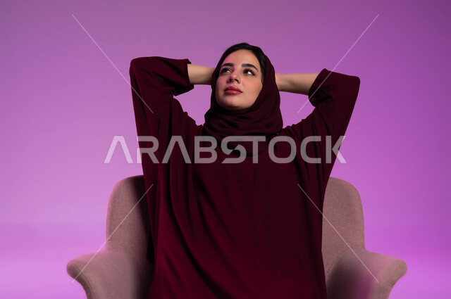 شرود الذهن و التمعن ،الشعور بالتعب و الخمول والحاجة إلى النوم, بورتريه مقرب لامرأة عربيه خليجيه سعوديه محجبه ترتدي العباءة الملونة تجلس على كرسي مريح تضع يديها وراء رأسها بتعابير التأمل، إيماءات الراحة والإسترخاء، النظر للأعلى إلى شيء ما، خلفية ملونه .