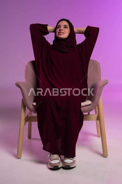 إيماءات الراحة والإسترخاء، الشعور بالتعب و الخمول والحاجة إلى النوم, بورتريه لامرأة عربيه خليجيه سعوديه محجبه ترتدي العباءة الملونة تجلس على كرسي مريح تضع يديها وراء رأسها بتعابير التأمل والسرور، النظر للأعلى إلى شيء ما، خلفية ملونه .