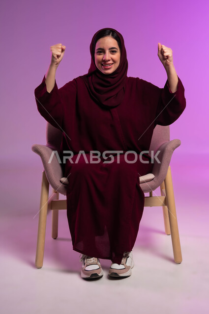 النظر الى الكاميرا بتعابير الفوز والكسب، ايماءات الفرح للانتهاء من انجاز شيء معين، مفهوم النجاح و تحقيق الاهداف، بورتريه لامرأة عربيه خليجيه سعوديه محجبه ترتدي العباءة الملونة تجلس على كرسي مريح وترفع يديها للأعلى بحركات التشجيع والحماس، خلفية ملونه .