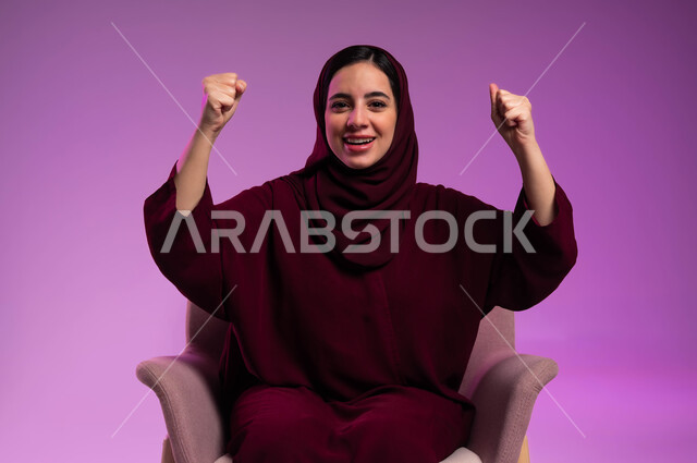 ايماءات الفرح للانتهاء من انجاز شيء معين، مفهوم النجاح و تحقيق الاهداف، النظر الى الكاميرا بتعابير الفوز والكسب، بورتريه لامرأة عربيه خليجيه سعوديه محجبه ترتدي العباءة الملونة تجلس على كرسي مريح وترفع يديها للأعلى بحركات التشجيع والحماس، خلفية ملونه .