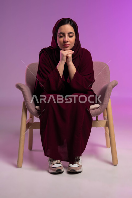الشعور بالنعاس والحاجه الى الراحة و النوم، بورتريه لامرأة عربيه خليجيه سعوديه محجبه ترتدي العباءة الملونة تجلس على كرسي مريح وتضع يديها تحت ذقنها بإيماءات الاسترخاء، اغماض العينين للتأمل وشرود الذهن، خلفية ملونة .