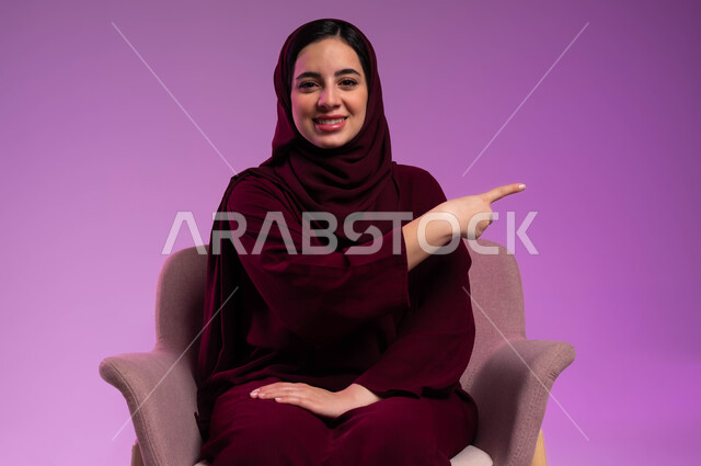 Pointing to something, looking at the camera with gestures of pleasure, expressions of confirmation and specifying a specific place, a portrait of a veiled Saudi Arabian Gulf woman, smiling, wearing a colorful abaya, sitting in a comfortable chair, raising her index finger to choose, a colored background.