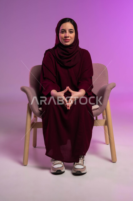 محاولة اخفاء التوتر وتخفيف الضغط النفسي، بورتريه لامرأة عربيه خليجية سعودية محجبة ترتدي العباءة الملونة تجلس على كرسي مريح و تقوم بملامسة أطراف الأصابع بعضها ببعض وتنظر إلى الكاميرا بإيماءات الثقة بالنفس، مفهوم الذكاء والإرادة القوية، خلفية ملونة .