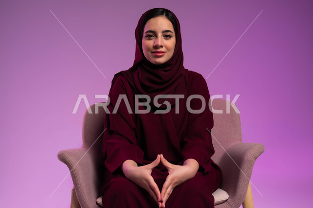 مفهوم الذكاء والإرادة القوية، بورتريه لامرأة عربيه خليجية سعودية محجبة ترتدي العباءة الملونة تجلس على كرسي مريح و تقوم بملامسة أطراف الأصابع بعضها ببعض وتنظر إلى الكاميرا بإيماءات الثقة بالنفس، محاولة اخفاء التوتر وتخفيف الضغط النفسي، خلفية ملونة .
