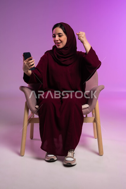 الفرح عند قراءة الأخبار السارة، تعابير السرور للفوز في مباراة كرة القدم ،الشعور بالحماس للنجاح وتحقيق الأهداف, بورتريه لامرأة عربيه خليجيه سعوديه محجبه مبتسمة ترتدي العباءة الملونة ترفع يدها للأعلى بايماءات الكسب، دور التقنية في توفير راحة أكبر للأفراد، خلفية ملونه .