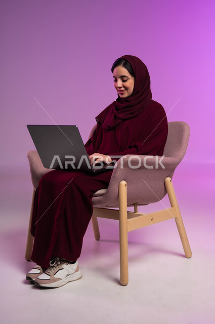 الدردشة والتواصل مع الاهل والأصدقاء، إدارة وتنظيم الاعمال، استخدام التقنيات الحديثة المتطورة والتكنولوجيا، مفهوم انجاز المهام اليومية، بورتريه لامرأة عربيه خليجيه سعوديه محجبه مبتسمة ترتدي العباءة الملونة تقوم بالعمل على جهاز الحاسوب المحمول، خلفية ملونه .