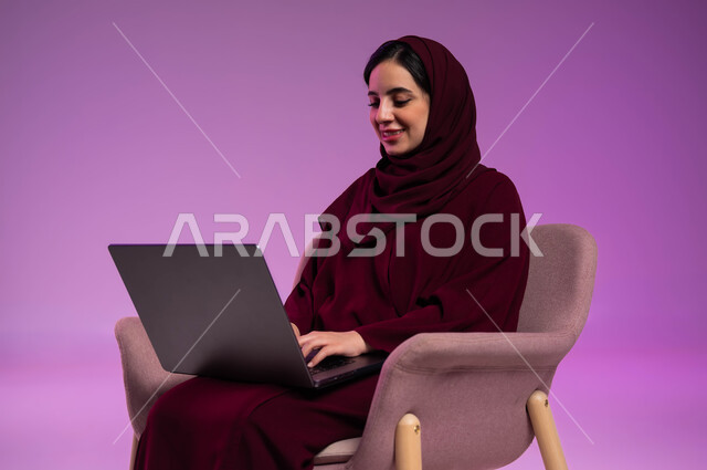 إدارة وتنظيم الاعمال، استخدام التقنيات الحديثة المتطورة والتكنولوجيا، مفهوم انجاز المهام اليومية، بورتريه لامرأة عربيه خليجيه سعوديه محجبه مبتسمة ترتدي العباءة الملونة تقوم بالعمل على جهاز الحاسوب المحمول، الدردشة والتواصل مع الاهل والأصدقاء، خلفية ملونه .