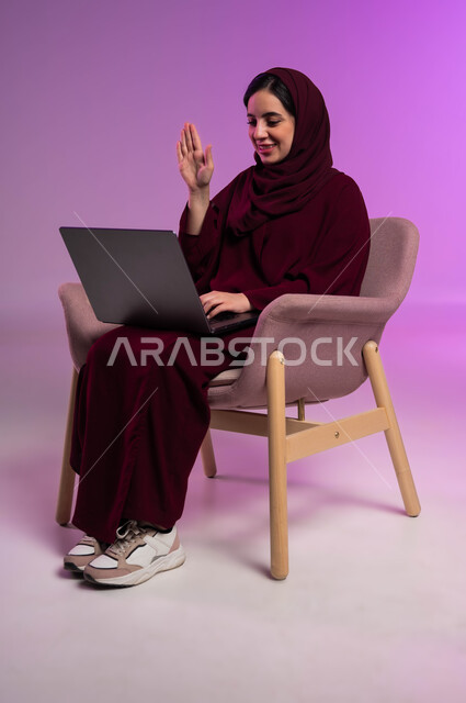 التواصل مع الأهل والاصدقاء، رفع اليد بتعابير القاء التحية والسلام، استخدام التقنيات الحديثة المتطورة ،بورتريه لامرأة عربيه خليجيه سعوديه محجبه ترتدي العباءة الملونة تجلس على كرسي مريح تقوم بإجراء مكالمة فيديو عبر جهاز الحاسوب المحمول، خلفية ملونه .