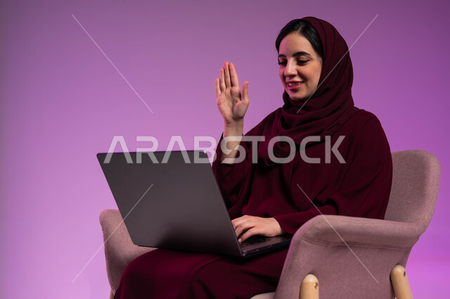 رفع اليد بتعابير القاء التحية والسلام، استخدام التقنيات الحديثة المتطورة ، التواصل مع الأهل والاصدقاء، بورتريه لامرأة عربيه خليجيه سعوديه محجبه ترتدي العباءة الملونة تجلس على كرسي مريح تقوم بإجراء مكالمة فيديو عبر جهاز الحاسوب المحمول، خلفية ملونه .