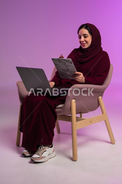 مجموعه من الأوراق الخاصة في العمل، انجاز المهام اليومية، بورتريه لامرأة عربيه خليجيه سعوديه محجبه مبتسمة ترتدي العباءة الملونة تقوم بالعمل على جهاز الحاسوب المحمول، تدوين وتسجيل الملاحظات، استخدام التقنيات الحديثة المتطورة والتكنولوجيا، خلفية ملونه .