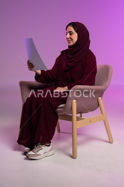ثقافة التعلم الذاتي, إنجاز الواجبات اليومية ،بورتريه لامرأة عربية سعودية خليجية محجبة ترتدي العباءة الملونة تجلس على كرسي مريح وتحمل بيديها ورقة بيضاء بايماءات السرور، الاستمتاع بقراءة القصص والروايات, خلفية ملونة .