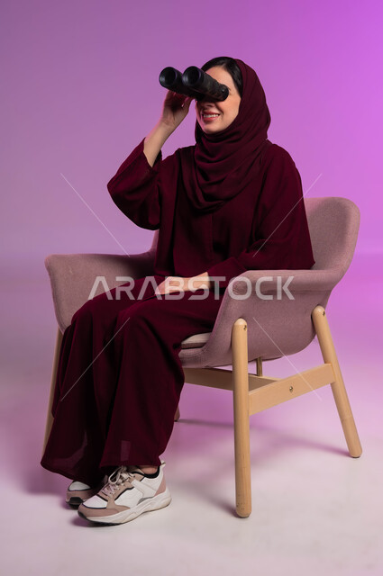 حب المغامرة و الاستكشاف، رؤية الاشياء البعيدة عبر التلسكوب، التخطيط المستقبلي للأهداف و الطموحات، بورتريه لامرأة عربية خليجية سعودية مبتسمة محجبة ترتدي العباءة الملونة تجلس على كرسي مريح و تنظر من خلال منظار ثنائي العينين, خلفية ملونة .