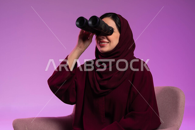 رؤية الاشياء البعيدة عبر التلسكوب، التخطيط المستقبلي للأهداف و الطموحات، حب الاستكشاف والمغامرة، بورتريه مقرب لامرأة عربية خليجية سعودية مبتسمة محجبة ترتدي العباءة الملونة تجلس على كرسي مريح و تنظر من خلال منظار ثنائي العينين, خلفية ملونة .