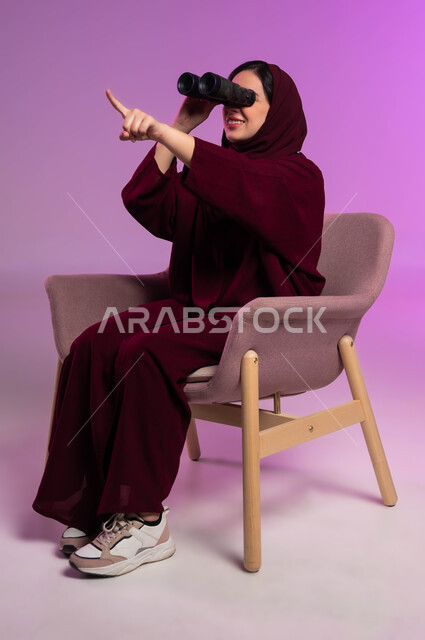 التخطيط المستقبلي للأهداف و الطموحات، الاشارة الى شيء ما، حب الاستكشاف والمغامرة، رؤية الاشياء البعيدة عبر التلسكوب، بورتريه لامرأة عربية خليجية سعودية مبتسمة محجبة ترتدي العباءة الملونة تجلس على كرسي مريح و تنظر من خلال منظار ثنائي العينين, خلفية ملونة .