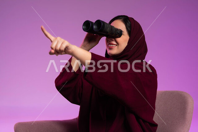 الاشارة الى شيء ما، حب الاستكشاف والمغامرة، رؤية الاشياء البعيدة عبر التلسكوب، بورتريه مقرب لامرأة عربية خليجية سعودية مبتسمة محجبة ترتدي العباءة الملونة تجلس على كرسي مريح و تنظر من خلال منظار ثنائي العينين, التخطيط المستقبلي للأهداف و الطموحات، خلفية ملونة .