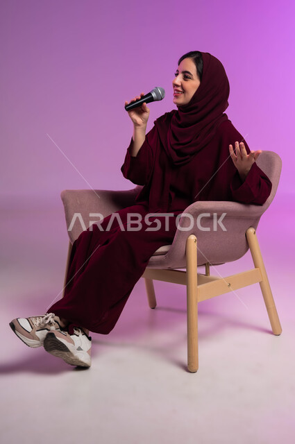 قضاء وقت ممتع بأداء العروض الصوتية، بدء التدريب الصوتي، ممارسة موهبة الغناء، مفهوم الفن وتأثيره الايجابي على الامرأة، بورتريه لشابة عربية خليجية سعودية محجبة ترتدي العباءة الملونة وتجلس على كرسي مريح تمسك بيدها المايكروفون بإيماءات السرور، خلفيه ملونة .
