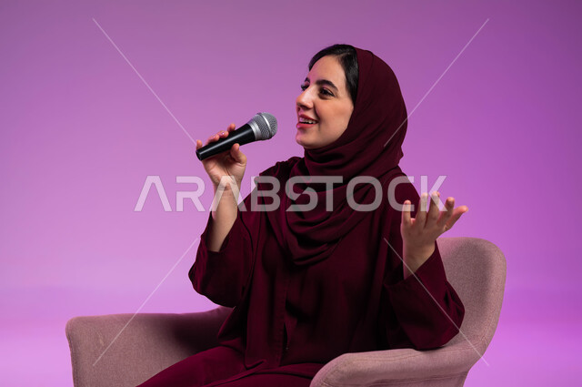 Practicing the talent of singing, spending an enjoyable time performing vocal performances, starting vocal training, the concept of art and its positive impact on women, a portrait of a veiled Saudi Gulf Arab young woman wearing a colorful abaya and sitting on a comfortable chair holding the microphone in her hand with happy gestures, a colorful background.