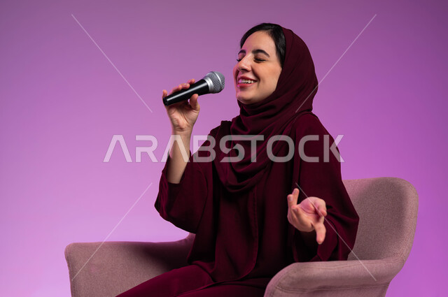 مفهوم الفن وتأثيره الايجابي على الامرأة، تقديم العروض الفنيه و الغنائيه والفقرات الترفيهية، بدء التدريب الصوتي، بورتريه لشابة عربية خليجية سعودية محجبة ترتدي العباءة الملونة وتجلس على كرسي مريح تمسك بيدها المايكروفون بإيماءات الاندماج بالغناء، خلفيه ملونة.