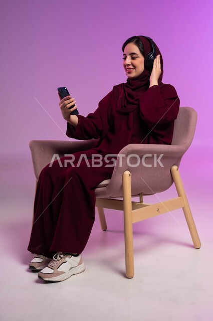الاستمتاع بسماع الموسيقا المفضلة، استخدام الأجهزة التقنية الحديثة المتطورة و التكنولوجيا، بورتريه لامرأة عربية سعودية خليجية محجبة مبتسمة ترتدي العباءة الملونة و سماعات الهيدفون تجلس على كرسي مريح وتنظر إلى الهاتف المحمول بايماءات السرور، خلفية ملونة .