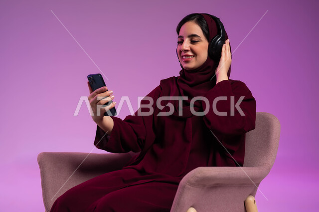 ايماءات السرور والاستمتاع بسماع الموسيقة المفضلة، استخدام الأجهزة التقنية الحديثة المتطورة و التكنولوجيا، بورتريه لامرأة عربية سعودية خليجية محجبة مبتسمة ترتدي العباءة الملونة و سماعات الهيدفون تجلس على كرسي مريح وتنظر إلى الهاتف المحمول، خلفية ملونة .