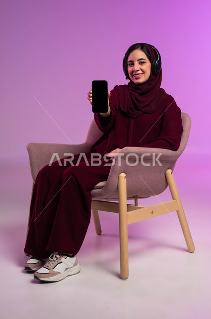 Using advanced modern technical devices and technology, a portrait of a smiling, veiled Saudi Arabian Gulf woman wearing a colorful abaya and headphones, sitting on a comfortable chair and looking at the camera with cypress gestures, showing a blank black screen on a mobile phone, a colored background.