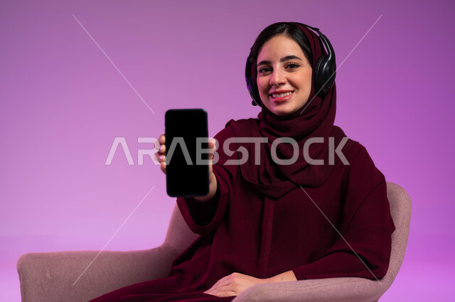 Displaying a blank black screen on a mobile phone, using technology and advanced modern technical devices, a portrait of a smiling veiled Saudi Arabian Gulf woman wearing a colorful abaya and headphones sitting on a comfortable chair and looking at the camera with cypress gestures, a colorful background.