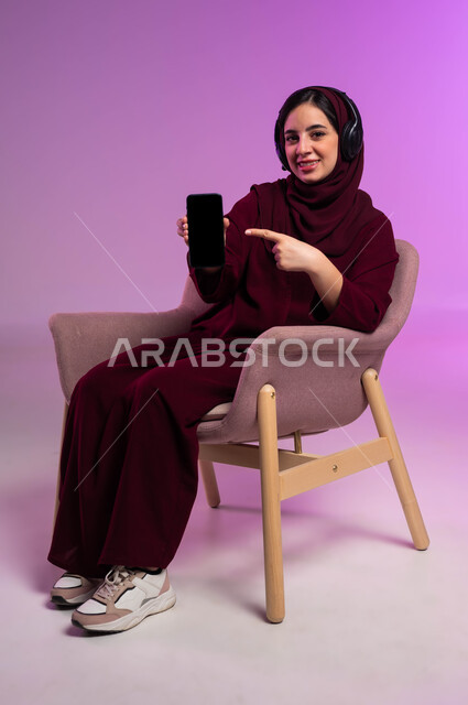 النظر إلى الكاميرا و الاشارة الى شاشه سوداء فارغه عبر الهاتف المحمول، استخدام التكنولوجيا و الأجهزة التقنية الحديثة المتطورة، بورتريه لامرأة عربية سعودية خليجية محجبة مبتسمة ترتدي العباءة الملونة و سماعات الهيدفون تجلس على كرسي مريح، خلفية ملونة .