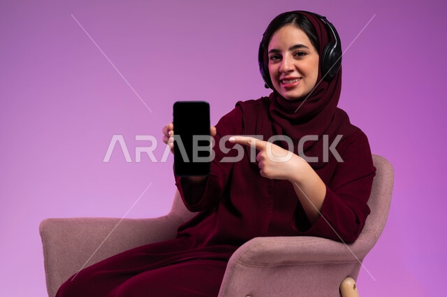 استخدام التكنولوجيا و الأجهزة التقنية الحديثة المتطورة، النظر إلى الكاميرا و الاشارة الى شاشه سوداء فارغه عبر الهاتف المحمول، بورتريه لامرأة عربية سعودية خليجية محجبة مبتسمة ترتدي العباءة الملونة و سماعات الهيدفون تجلس على كرسي مريح، خلفية ملونة .