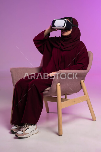 Technical and technological development, enjoying watching virtual reality movies and simulating the imaginary world, spending leisure time with recreational activities, portrait of a veiled Saudi Gulf Arab woman sitting in a comfortable chair, wearing a colorful abaya and 3D VR glasses, pink background