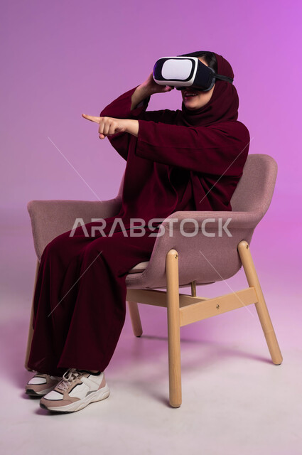 قضاء وقت ممتع بالأنشطة الترفيهية, الإشارة إلى عنصر ما بتفاعل واندماج, مشاهدة أفلام الواقع الافتراضي ومحاكاة العالم الخيالي, بورتريه لشابة عربية خليجية سعودية ترتدي العباءة الملونة والحجاب ونظارة في آر ثلاثية الأبعاد, خلفية وردية