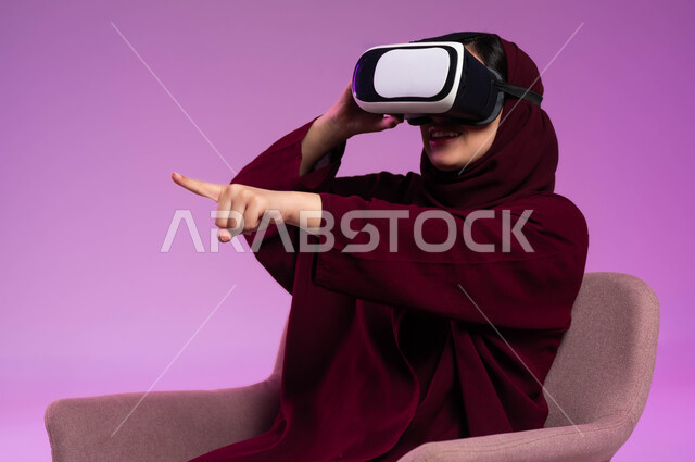 الإشارة إلى عنصر ما بتفاعل واندماج, مشاهدة أفلام الواقع الافتراضي ومحاكاة العالم الخيالي, قضاء وقت ممتع بالأنشطة الترفيهية, بورتريه لشابة عربية خليجية سعودية ترتدي العباءة الملونة والحجاب ونظارة في آر ثلاثية الأبعاد تشير إلى شيء ما, التطور التقني والتكنولوجي, خلفية وردية