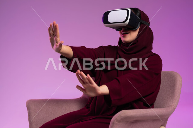 محاولة ابعاد وإيقاف شيء ما، التسلية والترفيه ومحاكاة العالم الخيالي، بورتريه لشابة عربية خليجية سعودية ترتدي العباءة الملونة ونظارة في آر ثلاثية الأبعاد ترفع يديها بحماس، التفاعل والاندماج مع الواقع الافتراضي، خلفية وردية