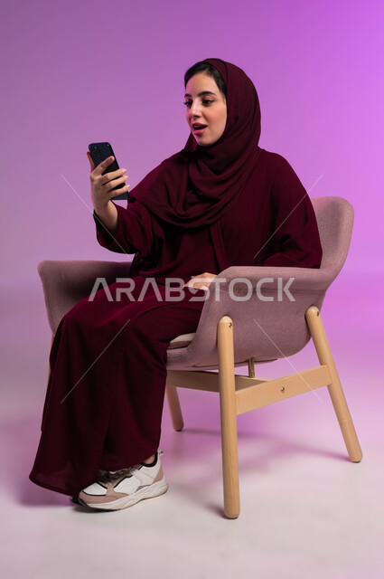 الاستمتاع في متابعه البرامج الترفيهيه المفضلة, إيماءات التفاجؤ والذهول, الاستغراب والانبهار والحماس, بورتريه لشابة عربية خليجية سعودية ترتدي العباءة الملونة والحجاب تستخدم تطبيقات الهاتف المحمول,  خلفية وردية