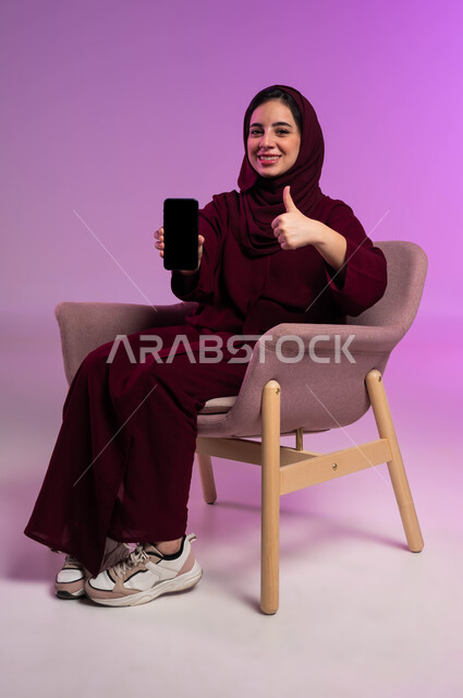 ايماءت الاعجاب والامتياز بالتطبيقات الالكترونية, استخدام الأجهزة التقنيه والتكنولوجيا الحديثه، جودة الخدمات وبرامج الهاتف المحمول، بورتريه لشابة عربية خليجية سعودية ترتدي العباءة الملونة والحجاب تمسك بيدها الجوال بشاشة سوداء فارغة, خلفية وردية