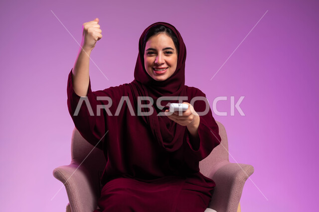 الاندماج والتفاعل بمشاهدة الأفلام السينمائية, الاستمتاع بقضاء أوقات مسلية, رفع اليد والنظر إلى الكاميرا بإيماءات السرور, بورتريه لشابة عربية خليجية سعودية مبتسمة ترتدي العباءة الملونة والحجاب تمسك بيدها جهاز التحكم عن بعد, خلفية وردية