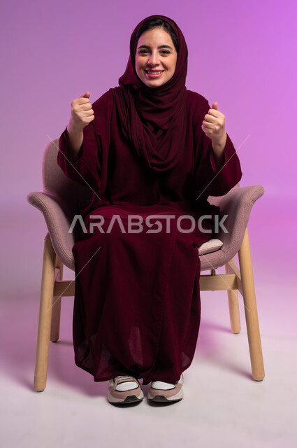 القوة والانتصار والتشجيع, النجاح والفوز وتحقيق الأهداف, بورتريه لشابة عربية خليجية سعودية مبتسمة ترتدي العباءة الملونة والحجاب ترفع يديها بإيماءات الحماس, الجلوس والنظر إلى الكاميرا بسرور, خلفية وردية