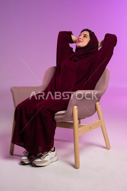 Sentiment de bonheur et de joie, expressions de contemplation, de pensée et de distraction, besoin de sommeil et de repos, portrait d'une jeune femme arabe du Golfe saoudienne portant une abaya colorée et un hijab assise sur une chaise et regardant quelque chose, corps sur toute la longueur portrait, fond rose