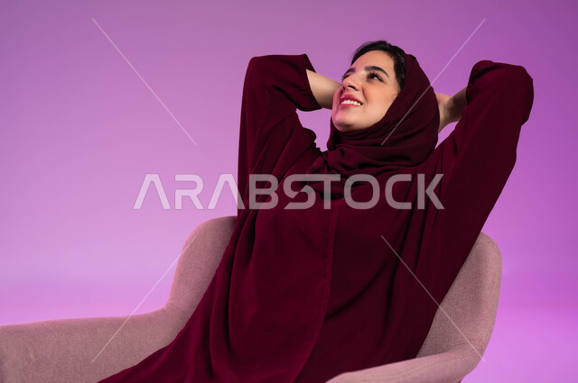 تعابير التأمل والتفكير وشرود الذهن، الشعور بالسعادة والفرح, الحاجة إلى النوم والراحة, بورتريه لشابة عربية خليجية سعودية ترتدي العباءة الملونة والحجاب تجلس على الكرسي وتنظر إلى شيء ما, خلفية وردية