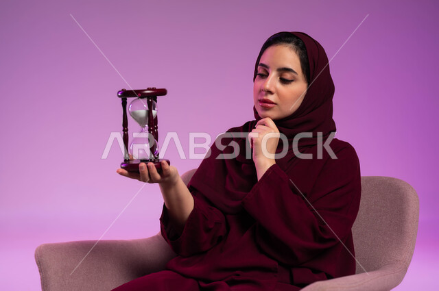 الجلوس بتعابير الملل والانتظار والسؤال, إدارة الأعمال وتنظيم الجدول الزمني, القيام بمراقبة توقيت الرمل, إدراك قيمة اللحظات الثمينة, بورتريه مقرب لشابة عربية خليجية سعودية ترتدي العباءة الملونة والحجاب تمسك بيدها الساعة الرملية الزجاجية (البنكام), خلفية وردية