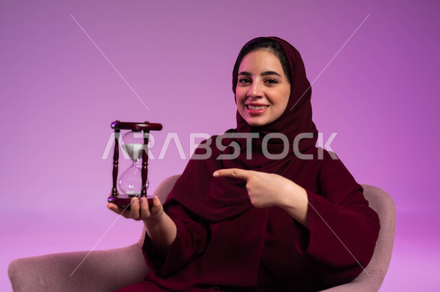 مفهوم إدارة الأعمال وتنظيم الجدول الزمني, القيام بمراقبة توقيت الرمل, إدراك قيمة اللحظات الثمينة, بورتريه مقرب لشابة عربية خليجية سعودية ترتدي العباءة الملونة والحجاب تمسك بيدها الساعة الرملية الزجاجية (البنكام), خلفية وردية