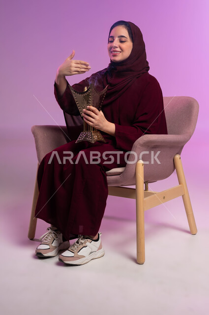 العادات والتقاليد السعودية, استخدام خشب العود بالأعياد والمناسبات, التطيب بالعود والبخور, بورتريه لشابة عربية خليجية سعودية ترتدي العباءة الملونة والحجاب تقوم بالتعطر من حامل البخور, خلفية وردية