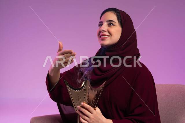 العادات والتقاليد السعودية, استخدام خشب العود بالأعياد والمناسبات, التطيب بالعود والبخور, بورتريه لشابة عربية خليجية سعودية ترتدي العباءة الملونة والحجاب تنظر إلى شيء ما وتقوم بالتعطر من حامل البخور, خلفية وردية