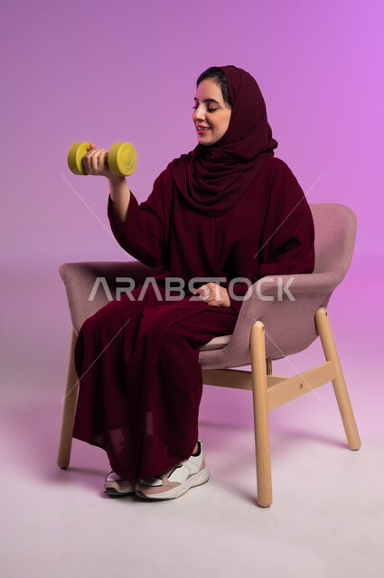 اللياقة والليونة والرشاقة البدنية, ممارسة الأنشطة الرياضية, بورتريه مقرب لسيدة عربية خليجية سعودية محجبة مبتسمة ترتدي العباءة الملونة تحمل بيدها الاوزان الثقيلة الحديدية ( الدمبلز) تنظر إليها, اتباع أسلوب حياة صحي, خلفية بنفسجية