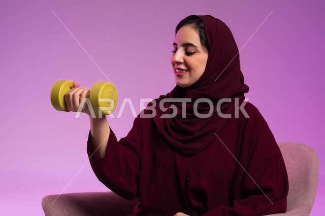 مفهوم اللياقة والليونة والرشاقة البدنية, ممارسة الأنشطة الرياضية, بورتريه مقرب لسيدة عربية خليجية سعودية محجبة مبتسمة ترتدي العباءة الملونة تحمل بيدها الاوزان الثقيلة الحديدية ( الدمبلز) تنظر إليها, اتباع أسلوب حياة صحي, خلفية بنفسجية