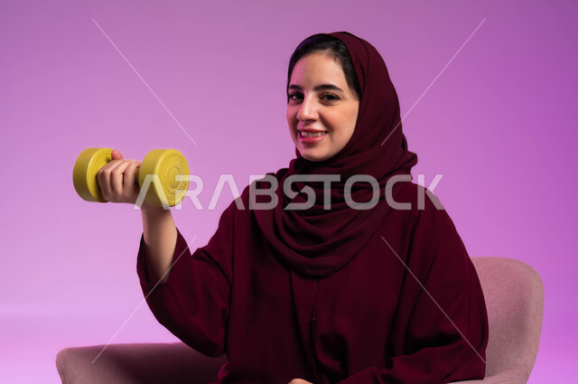 مفهوم اللياقة والليونة والرشاقة البدنية, اتباع أسلوب حياة صحي, ممارسة الأنشطة الرياضية, بورتريه مقرب لسيدة عربية خليجية سعودية محجبة مبتسمة ترتدي العباءة الملونة تحمل بيدها الاوزان الثقيلة الحديدية ( الدمبلز) تنظر إلى الكاميرا بإيماءات الثقة, خلفية بنفسجية