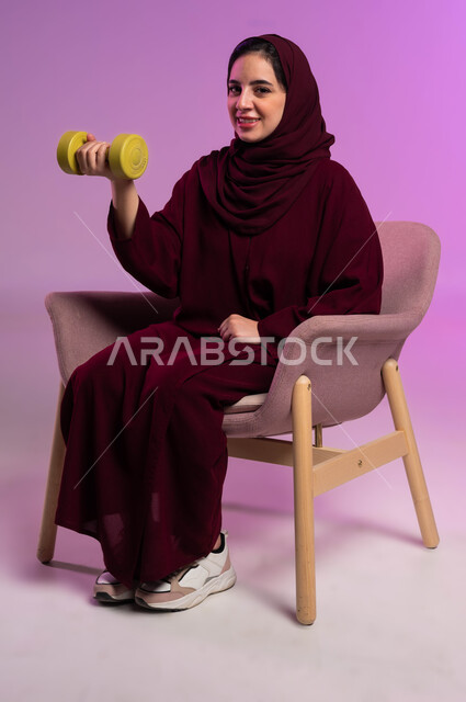 Following a healthy lifestyle, practicing sports activities, a portrait of a veiled Saudi Arabian Gulf woman, smiling, wearing a colorful abaya, holding heavy iron weights (dumbbells) in her hand, looking at the camera with confident gestures, the concept of fitness, flexibility and physical agility, purple backgroun